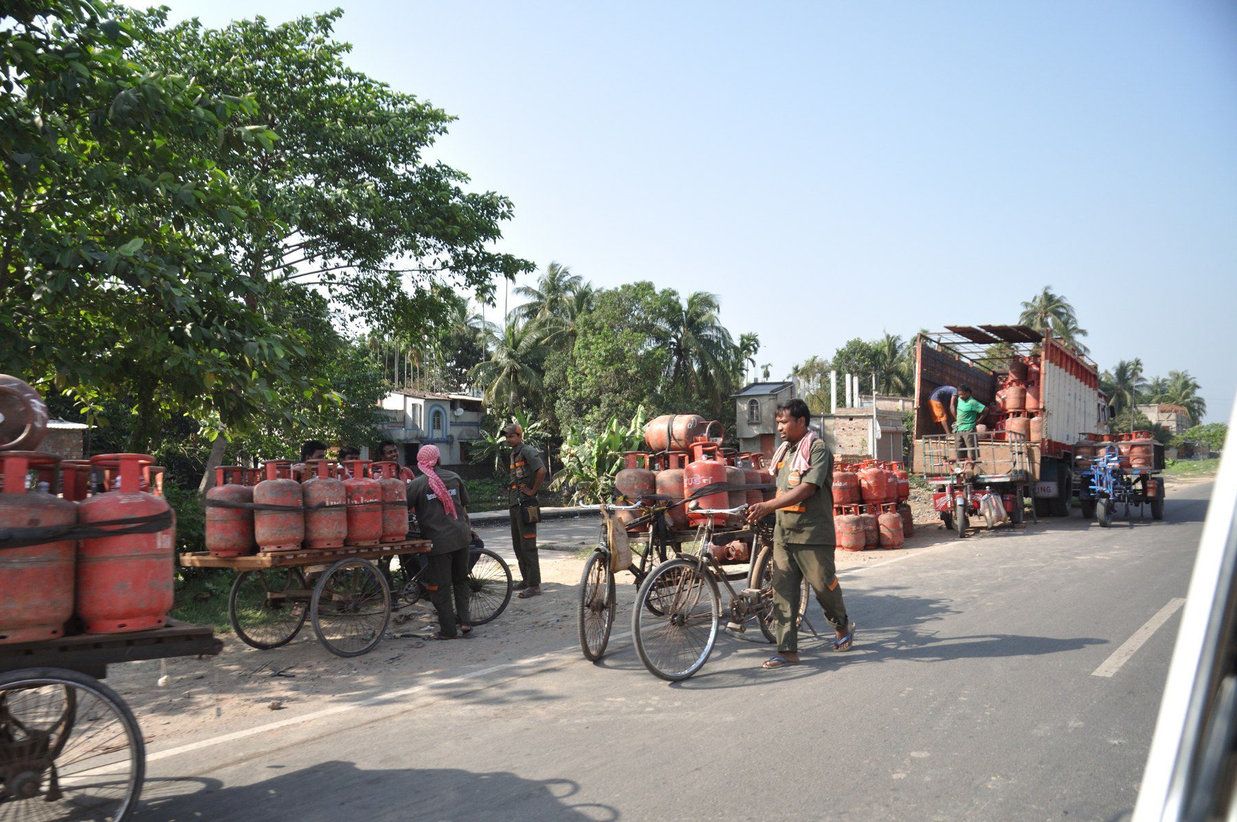 Delivery workers loading domestic LPG cylinders onto cycle carts beside a truck on a roadside