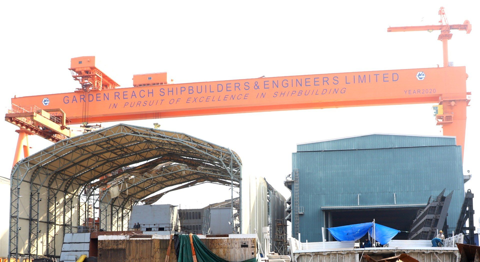 Orange gantry crane bearing the Garden Reach Shipbuilders and Engineers name over shipbuilding sheds in Kolkata