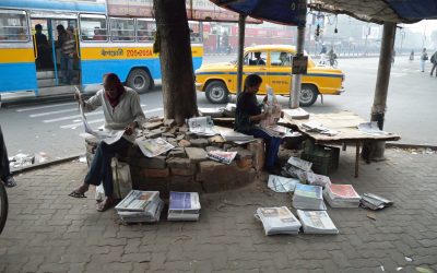 People reading newspapers at a roadside news stand with stacks of Indian dailies on the pavement